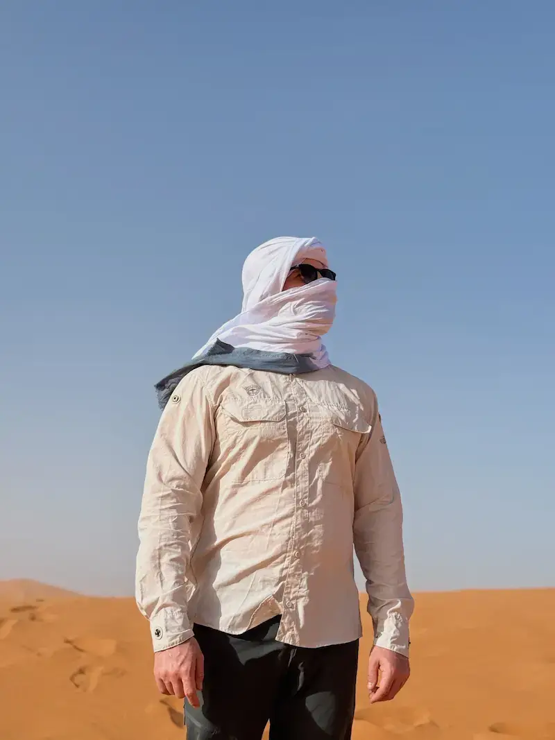 A traveler standing against a backdrop of orange sand dunes and a clear blue sky in the Sahara Desert, wearing recommended sun-protective clothing including a long-sleeved shirt, sunglasses, and a traditional white Berber turban (chèche) completely wrapping his head and face